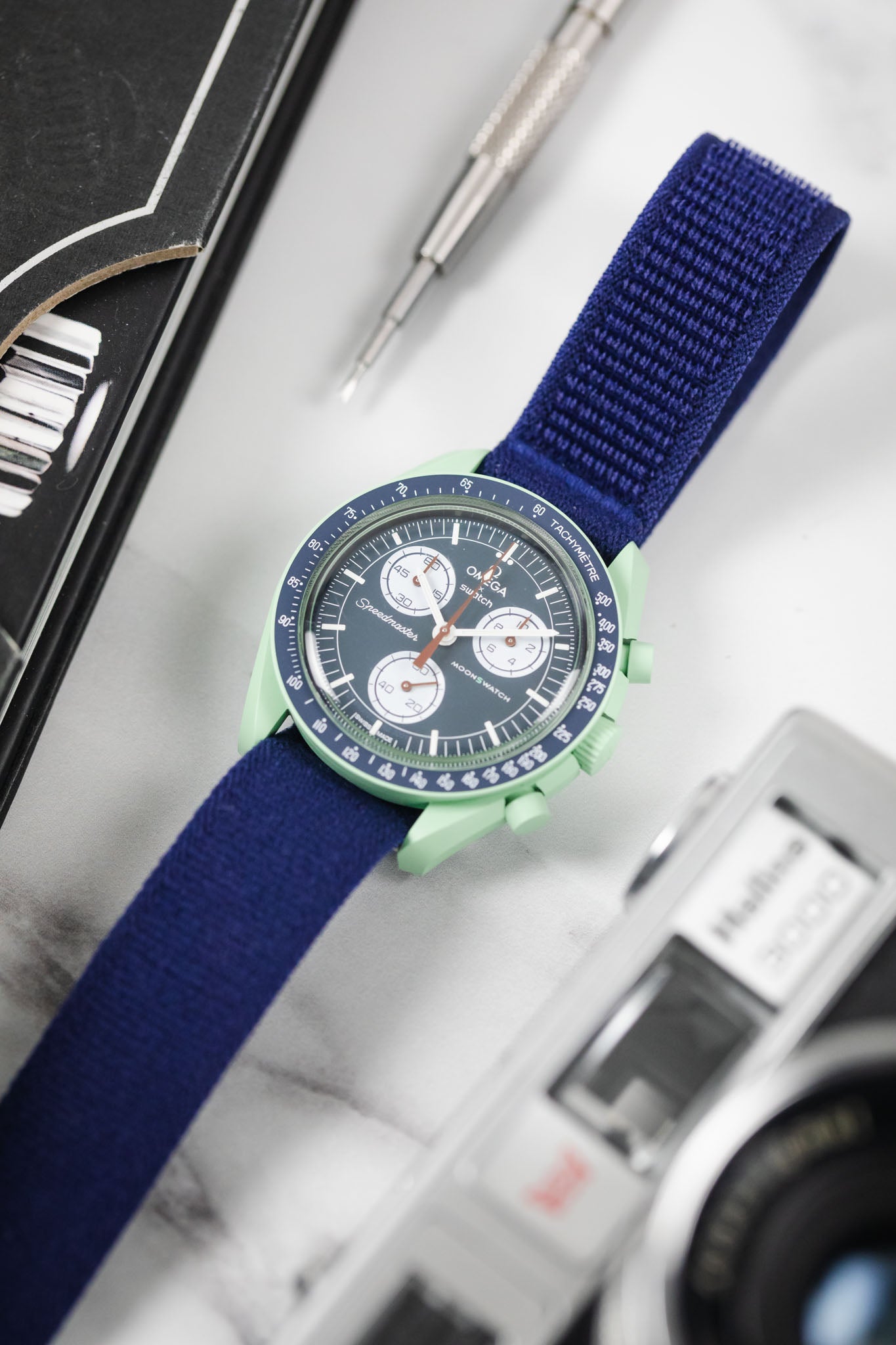 OMEGA x Swatch Mission on Earth watch on a vibrant blue textile watch strap, with vintage camera and tool.