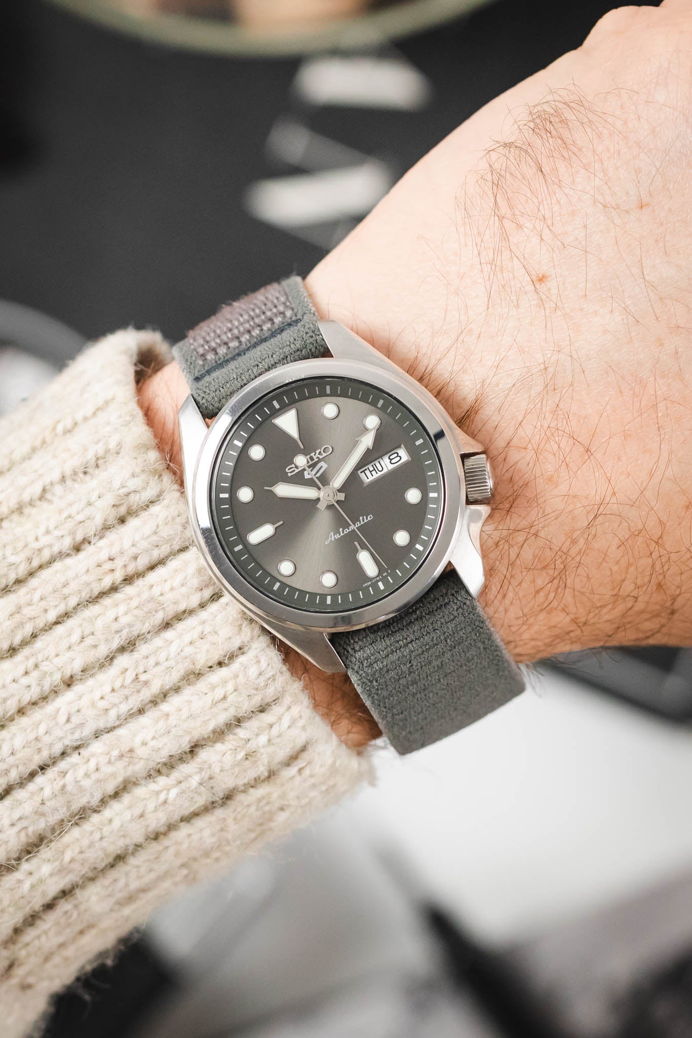 Seiko 5 Sports watch with a grey dial on a HIRSCH Vocem Grey strap, worn on a wrist over a knit sweater.