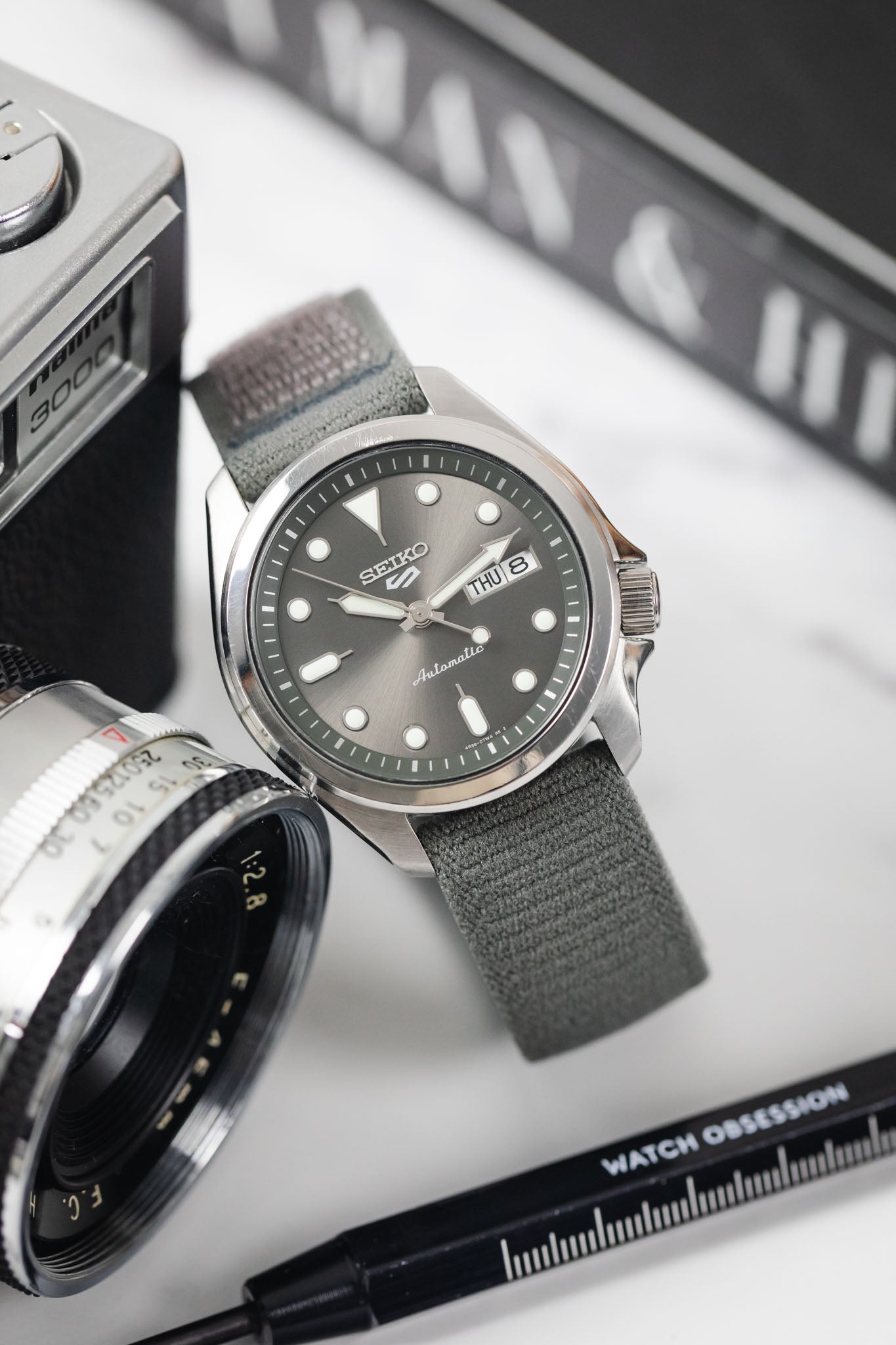 Close-up of Seiko 5 Sports automatic grey watch on a HIRSCH Vocem Grey strap with surrounding accessories.