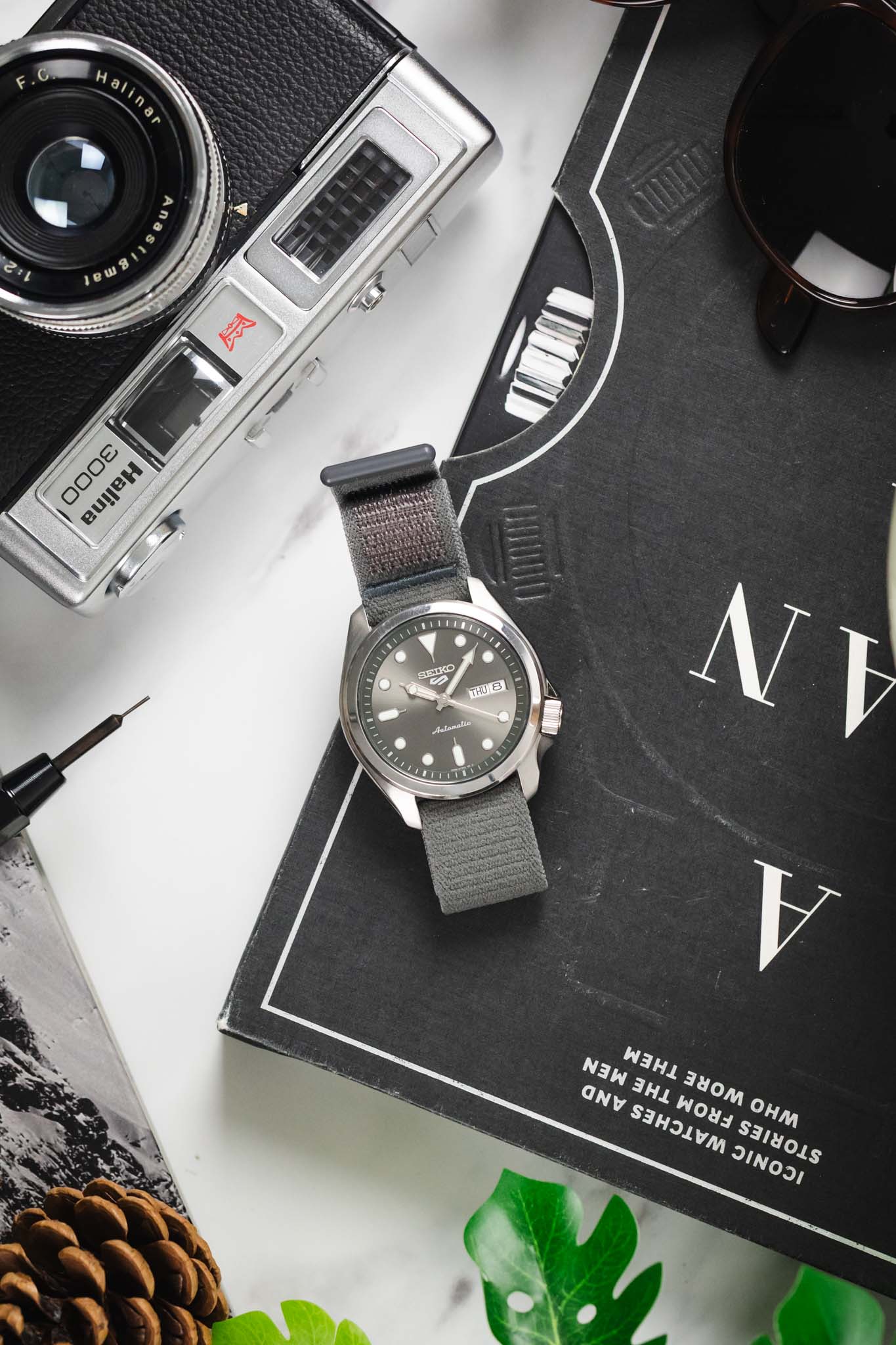 Seiko 5 Sports grey watch on a HIRSCH Vocem Grey strap, laid flat with a vintage camera and sunglasses.