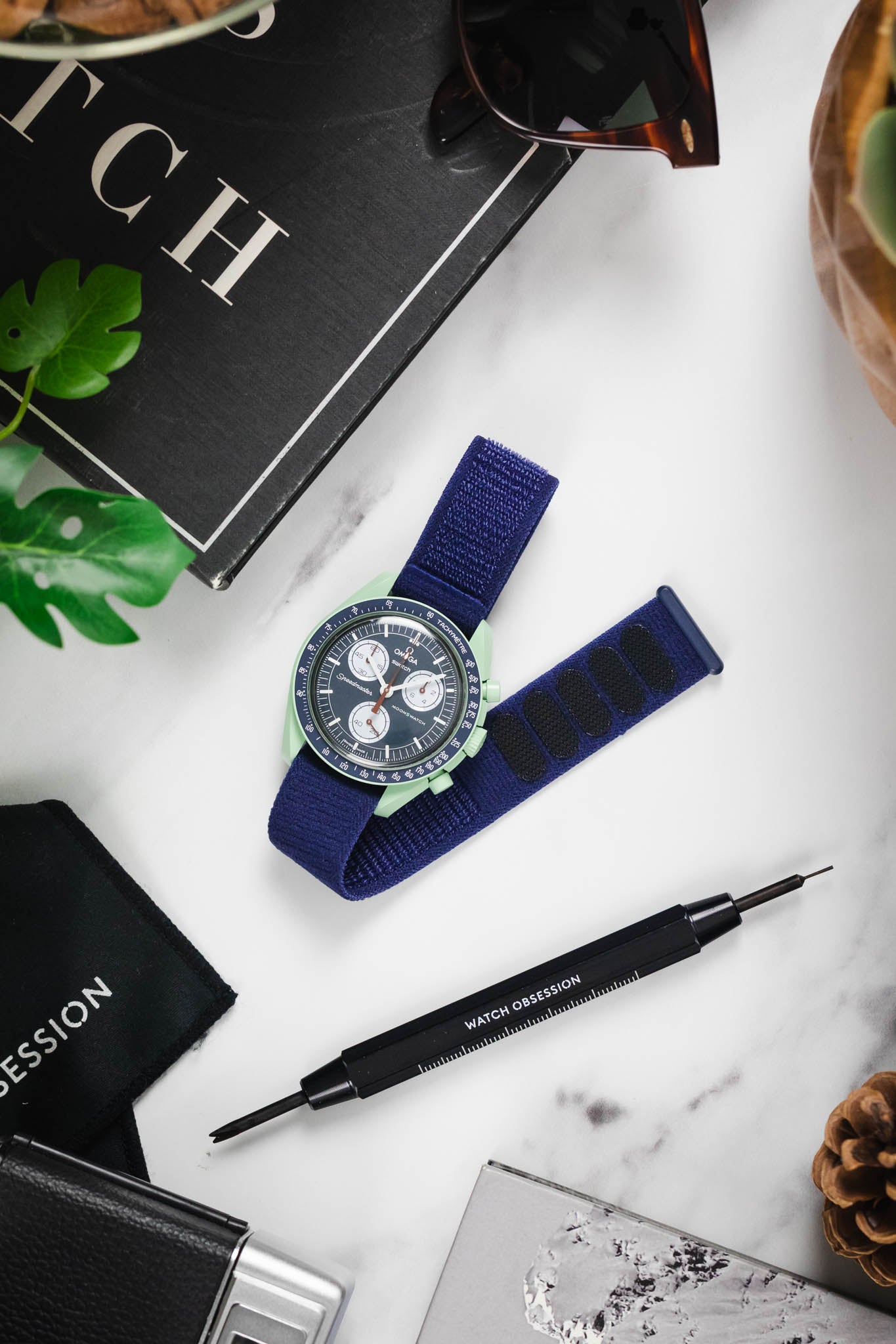 Flat lay of OMEGA x Swatch Mission on Earth watch on a blue textile strap with various watch accessories.