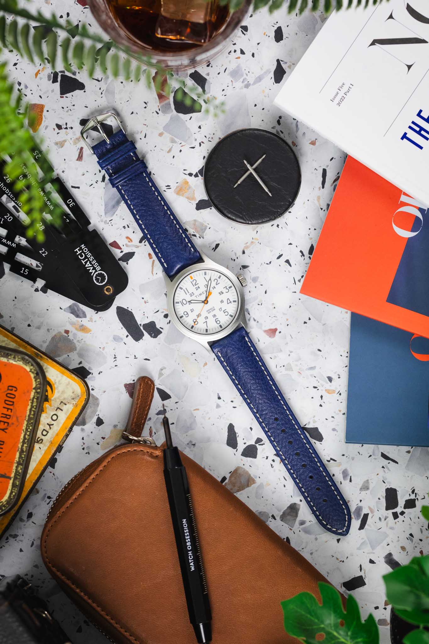 Buy the Hirsch Boston Blue leather watch strap. Shown with a white dial field watch on a terrazzo background.