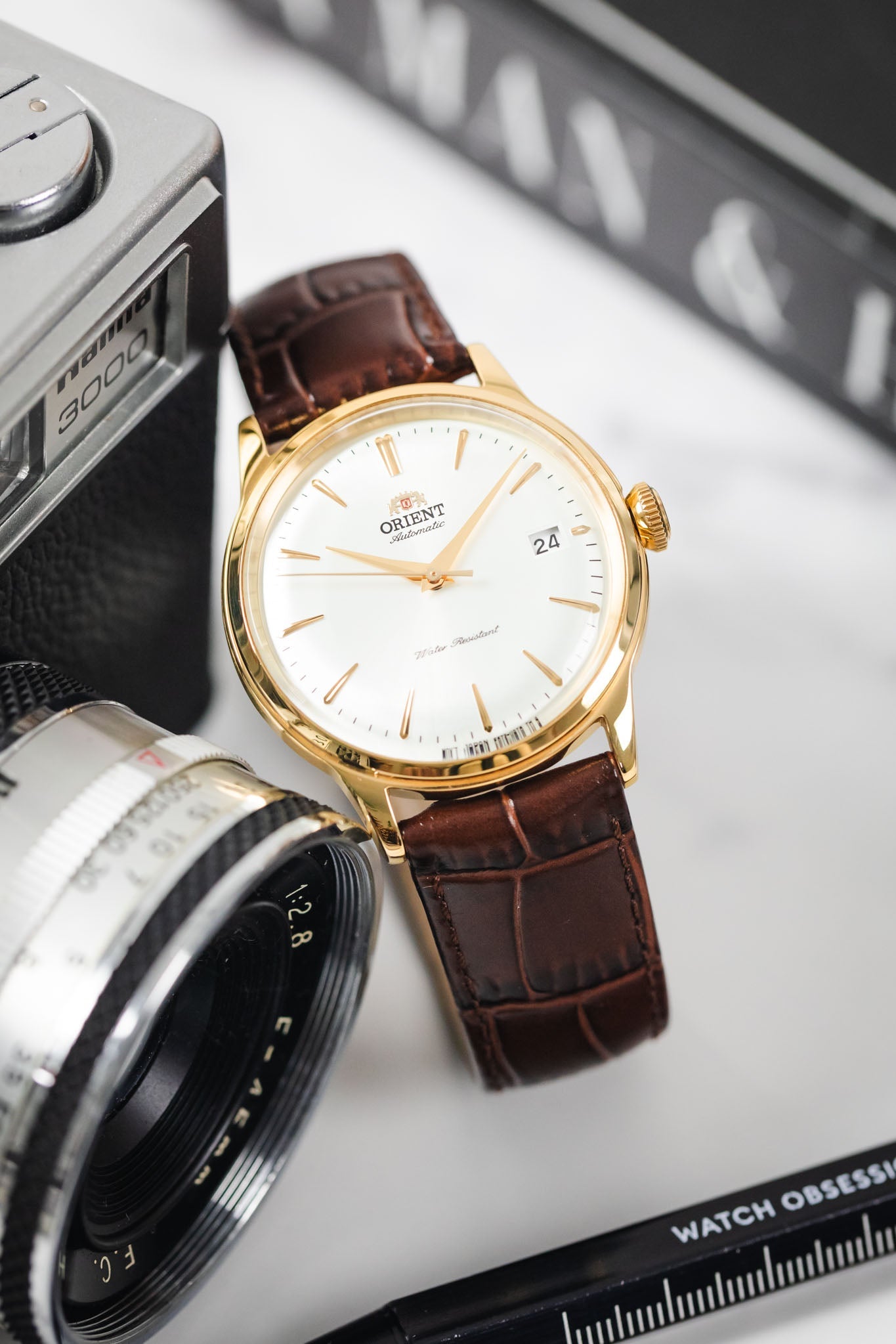 Gold-case Orient automatic watch on a brilliant brown Louisianalook strap, near a camera and book.