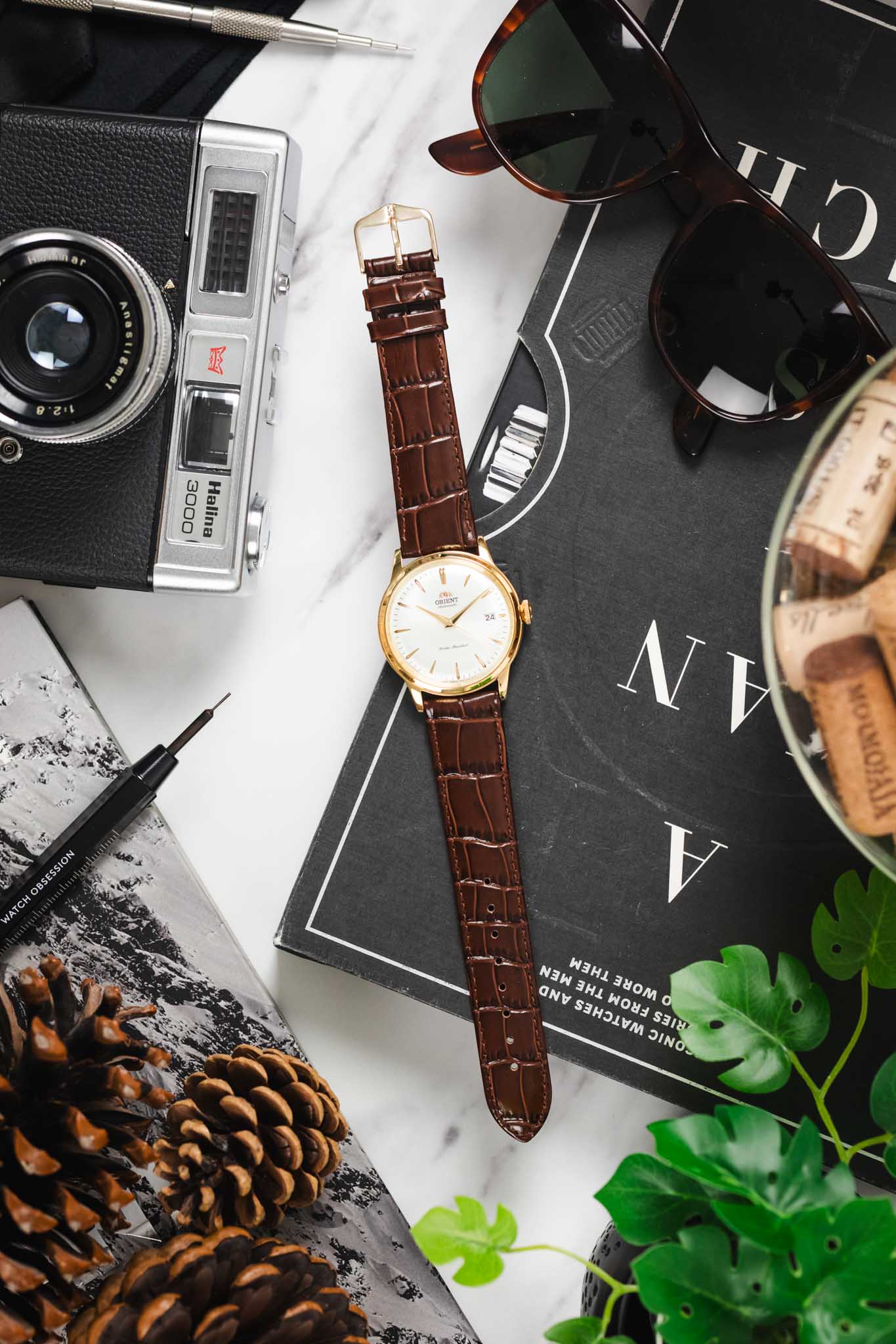 Flat lay of a gold-case Orient watch on a brown Louisianalook strap, styled with a tool and sunglasses.