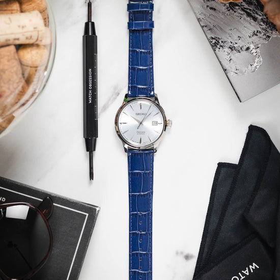 Flat lay of a Seiko Presage watch on a blue Louisianalook strap, styled with a spring bar tool and other gear.