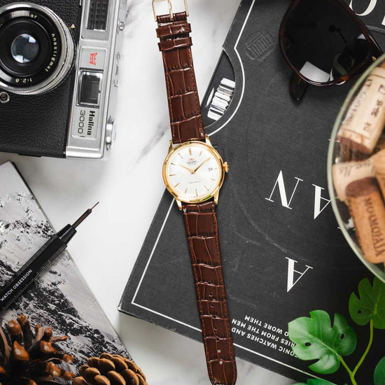 Flat lay of a gold-case Orient watch on a brown Louisianalook strap, styled with a tool and sunglasses.
