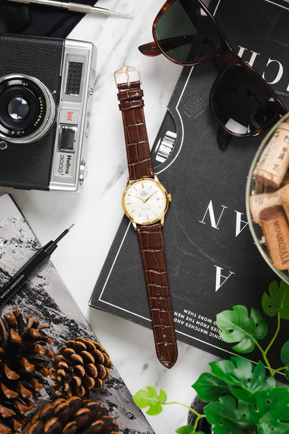 Flat lay of a gold-case Orient watch on a brown Louisianalook strap, styled with a tool and sunglasses.